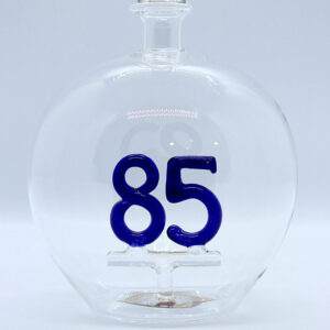 Eine leere bauchige Flasche aus Glas mit einer aus Glas geblasenen Zahl 85 im Inneren.