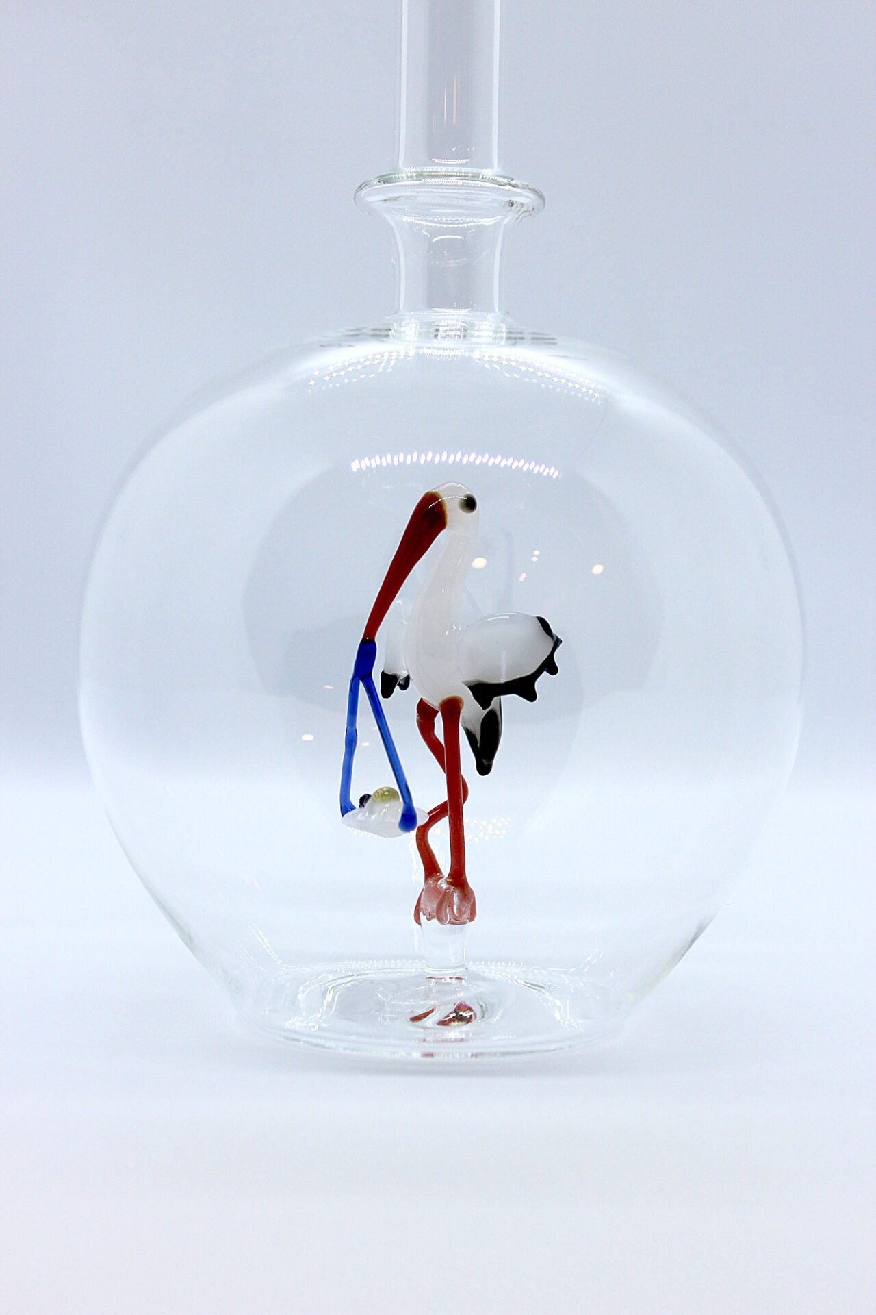 Eine leere bauchige Flasche aus Glas mit einem aus Glas geblasenen Storch mit Baby im Inneren.